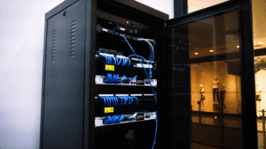 Imagem de um rack de servidores com porta de vidro aberta, exibindo a nova infraestrutura de rede instalada pela Whido para a Los Grandes (LOS). A estrutura conta com cabeamento organizado, switches de rede modernos e equipamentos de conectividade de alto desempenho. Ao fundo, é possível ver parte de um espaço interno moderno com iluminação controlada e manequins decorativos.