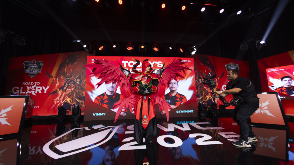Cosplayer com armadura vermelha e asas gigantes no palco do SWC Copa Americas segurando troféu, durante evento com iluminação e cenografia.